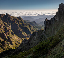 Fototapeta premium Warm morning light highlights rugged green mountains on Madeira Island, Portugal, as they rise above a vast sea of clouds, seen from Pedra Rija viewpoint under a clear blue sky