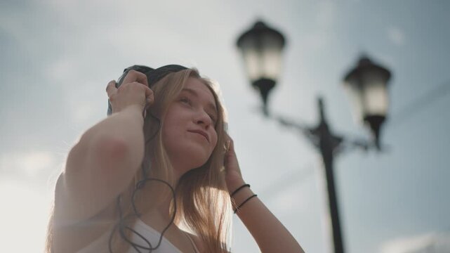 Sunlit street portrait white teen headphones, golden backlight and lamppost silhouette, braided hair catching breeze, adjusting overear headset while smiling toward sky, scenes show street dancer