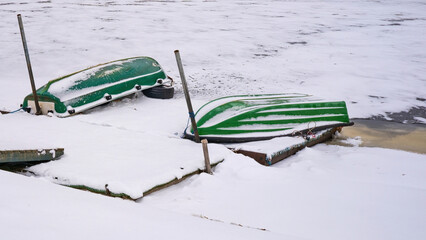 Upside-down green boats covered with snow on a frozen shoreline in winter. Abandoned small vessels on icy water, cold season landscape, off-season boating concept, minimal winter scene 