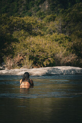 Mujer de espaldas ba&ntilde;&aacute;ndose en un r&iacute;o cristalino rodeado de naturaleza.