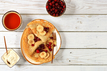 Delicious Russian pancakes with jam, soft cottage cheese, bananas, cranberries, and honey on a marble table. Healthy breakfast made from natural ingredients, Maslenitsa concept, natural products