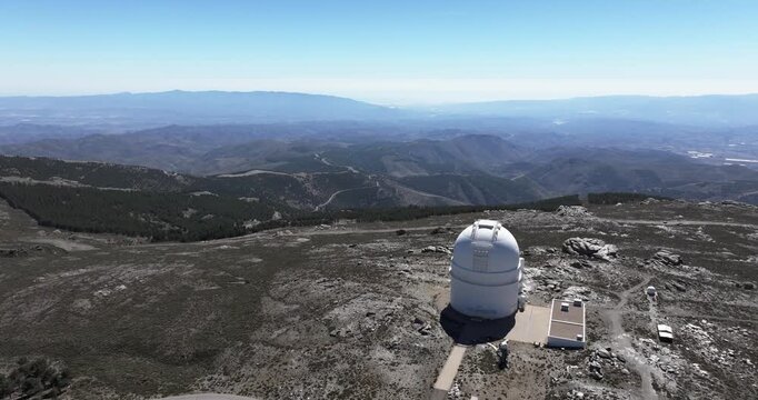 Wide drone aerial of Calar Alto Observatory dome in Tabernas Spain, barren rocky terrain and distant mountains under clear blue sky
