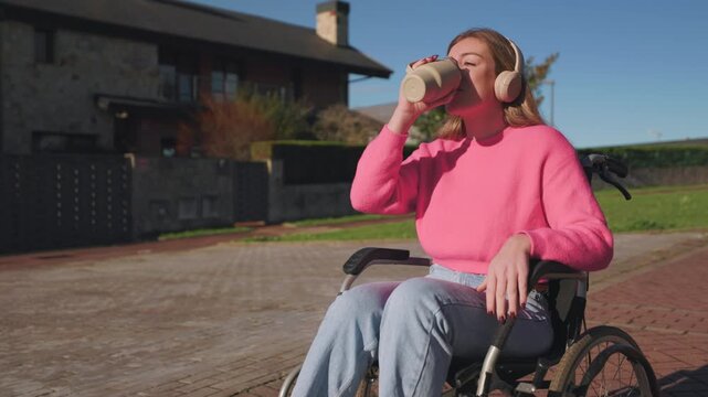 Happy young woman with a disability sitting in a wheelchair while listening to music with headphones and drinking from a reusable coffee cup outdoors on a sunny day in the neighborhood