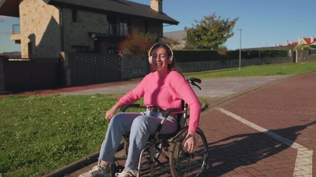 Joyful paraplegic woman wearing headphones and dancing with open arms while riding her wheelchair outdoors, embracing life with positivity and celebrating freedom on a sunny day