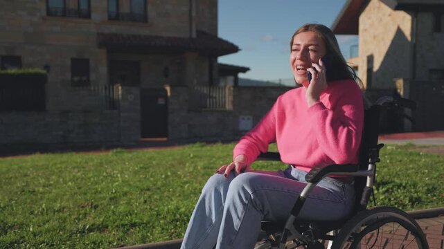 Cheerful young paraplegic woman with a disability sitting in a wheelchair while having a pleasant conversation on her smartphone in a residential area on a sunny day