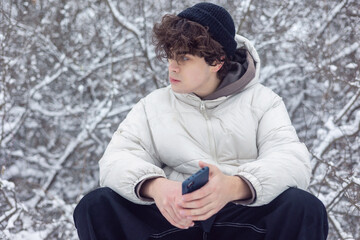 teenage boy sitting outdoors in a snowy forest while holding a smartphone during a winter walk. modern youth lifestyle. winter season, everyday life, and technology.