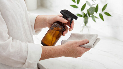 Woman's hands holding an amber glass spray bottle and a microfiber cloth for eco-friendly cleaning in a bright kitchen.