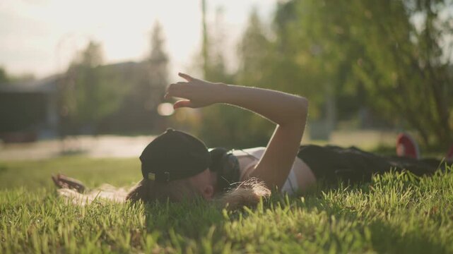 white female dancer lying in sunlit grass, cap and red sneakers visible street performer taking break, choreographer testing floor rolls, model posing for urban photoshoot, close lowangle shots