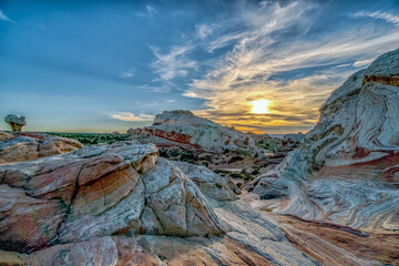 A beautiful sunset over a rocky landscape. The sky is filled with clouds and the sun is setting in the distance. The rocks are white and red, creating a striking contrast against the blue sky