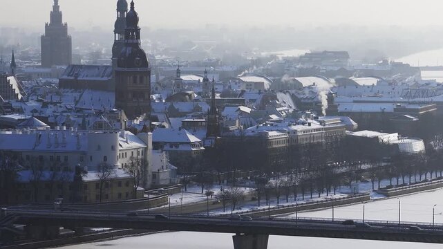 Aerial view over Riga, Latvia, showing Old Town, Vansu Bridge cable stays, the frozen Daugava, church towers, arched railway bridge, cars, steam, and soft backlight haze.