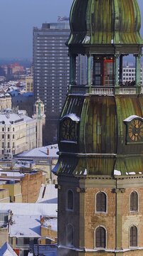 Aerial view over Riga, Latvia shows a church tower with green copper dome and clocks, people on balcony, Nativity Cathedral domes, Radisson high rise, and snowy roofs.