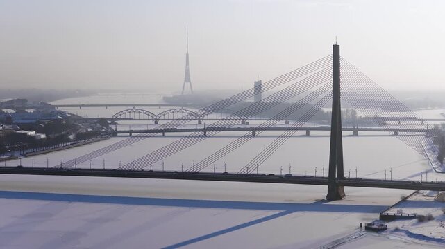 Aerial view of frozen Daugava in Riga with Vansu Bridge cables casting long shadows on ice, slow drone motion, sparse traffic, Riga TV Tower spire in haze.