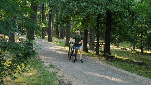 A woman walks with a bicycle through a shaded forest park path on a summer day. The moment conveys relaxation, motion and a natural outdoor routine.