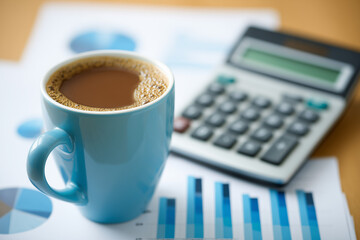 blue coffee mug filled with coffee on printed charts and graphs beside calculator on wooden desk