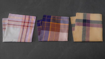 Collection of colorful plaid and checkered vintage cotton handkerchiefs neatly folded and arranged on a dark grey surface.