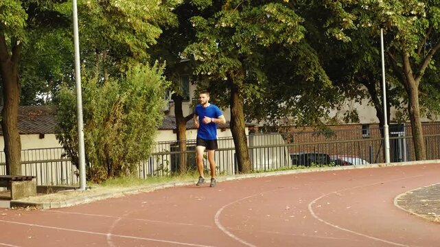 A man in sportswear jogs along a stadium track surrounded by trees and stops as sudden leg pain forces him to slow down. He begins to limp and heads toward a nearby bench to sit and rest