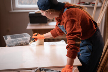 Professional woman carpenter painting wooden furniture part with brush and beige oil paint in craft workshop