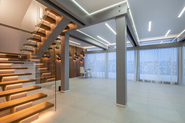 A room with a modern design features a wooden staircase, bright lighting, and large windows...