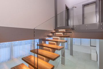 The scene shows a modern staircase featuring wooden steps with a glass railing inside a bright...
