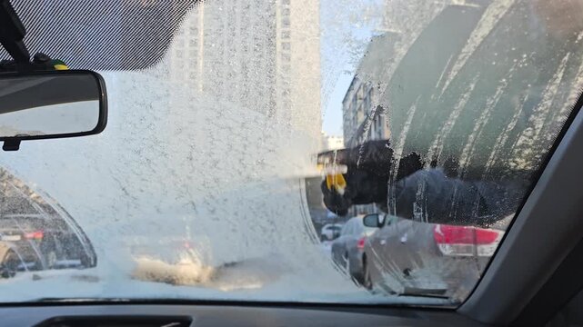 View from the interior of an automobile to a man in warm clothes cleaning the windshield of a car from snow, using an ice scraper. Winter car maintenance. 