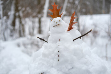 A tiny snowman figure decorated with dry brown leaves and twig arms resting on a snowy stump in the woods.
