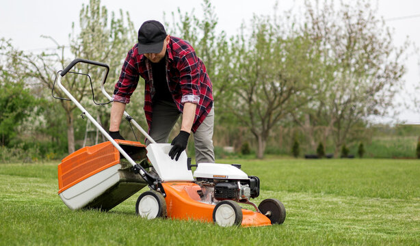Man in casual clothes takes out bag with cut grass of lawn mower after mowing lawn with at own backyard. Husband takes care of garden. Modern gasoline garden equipment. Landscaping work at home