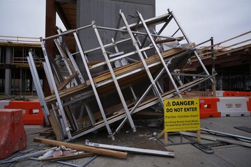 A construction site shows a collapsed scaffolding next to a road. A warning sign instructs people not to enter the area while workers assess the situation.