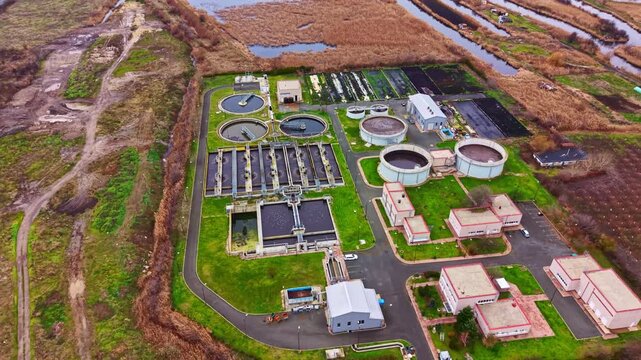 A water treatment facility is seen from above. It has several round and rectangular tanks, buildings, and open areas. The surrounding land has grass and patches of water during the day.