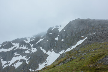 Minimal alpine view from grassy pass above abyss to sharp rocky ridge with snow in rainy haze. Minimalist landscape in gloomy weather. Vertical rock wall with snowfields under gray sky in rain time.