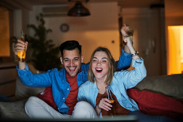 Portrait of an excited young couple watching tv and screaming while drinking beer and cheering for their national soccer team victory