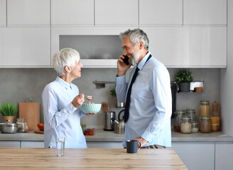 Portrait of a mid aged mature woman and man  gettyng ready for work using a smartphone and eating breakfast oatmeal in a bowl standing in kitchen home. Overworked, busy, stress, urgency, multitasking,