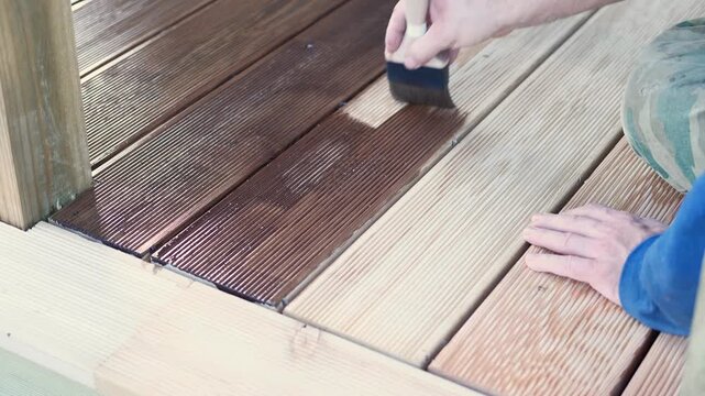 Brown oil for protection of wood by staining. Painting a larch for protecting and pests. Woodworker applying finish to terrace deck board. Repairman varnishing with brush wooden plank floor surface.