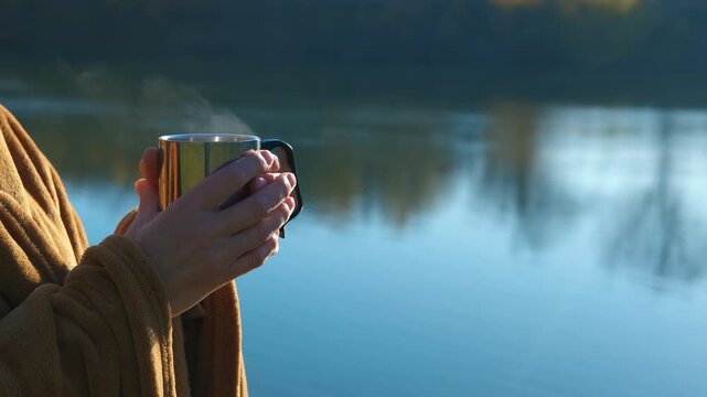 Woman holding a hot metal cup of tea by the lake. Female hands in a cozy bathrobe holding a warm metal mug with a steaming drink, enjoying a morning by the lake with calm water reflecting the nature