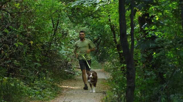 A man runs along a narrow forest path with his Australian Shepherd dog pulling ahead on a canicross belt. The concept highlights teamwork, speed, and energetic outdoor movement with pet