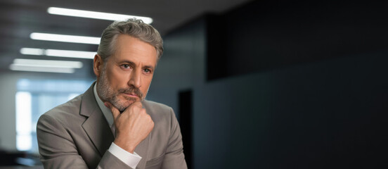 Mature businessman in thought with hand on chin in modern office with cool lighting for corporate use.