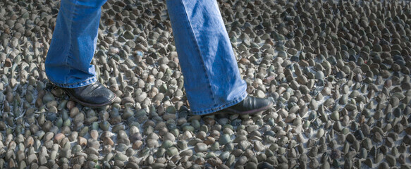 A person wearing jeans and boots walks over a path made of smooth pebbles. The composition focuses on the lower part of the legs and the texture of the stones.