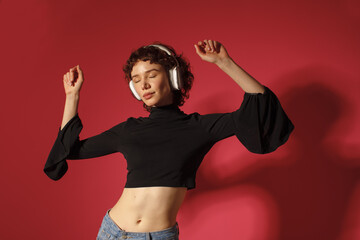 Young woman dancing and listening to music with headphones on red background. Freedom, joy,...