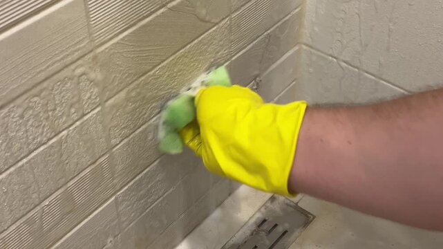 60 fps video close up. Male hand in yellow latex glove cleans wall with ceramic tiles with coating, rust, dirt in shower cabin of home bath room with sponge. Concept of housework, hygiene, wash