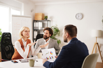 Group of professionals gathers around a table in a bright office room. They discuss ideas and share insights while reviewing charts and graphs to plan for future success.