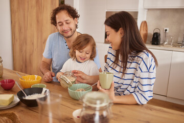 A cheerful family is gathered around the kitchen table preparing breakfast. Parents and child are smiling, enjoying each other's company and sharing food.