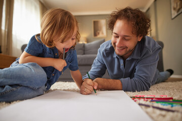 A father and his young child are lying on a soft carpet, coloring and drawing with various pencils. The cozy living room has warm light and a comfortable sofa.