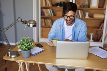 A man dressed casually smiles while holding a coffee mug at his wooden desk, engaging with his laptop and surrounded by books and office supplies.