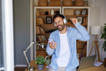 In a bright home office, a man wears headphones, smiles, and dances while checking his smartphone. The workspace features plants and shelves filled with books, creating a relaxing atmosphere.