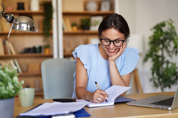 A woman sits at a wooden desk in her home office, writing in a notebook with a smile. The cozy space features shelves filled with books and decorative plants, creating an inspiring environment.