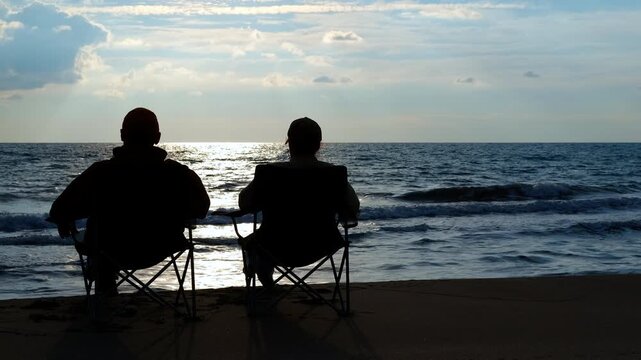 Two silhouetted friends sitting on a beach watching the sunset. Two male friends sitting on camping chairs on a sandy beach, their silhouettes outlined by the sunset reflecting on the calm sea