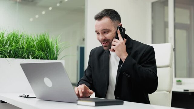 Businessman takes a phone call while working on a laptop, showing focused expressions and gestures in a modern office with greenery. Concept productivity, concentration, modern work