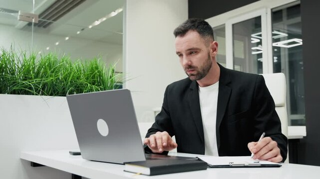 Professional man in a black suit focuses on a laptop, typing and taking notes. Productivity and engagement in a modern office with greenery. Concept work, concentration, modern office