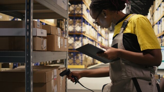 African american e-commerce worker scanning shipping labels numbers, registering awb and shipment details for the inventory records. Young man handling pallets distribution operations. Camera A.