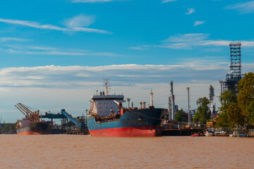 Cargo ships docked at an industrial port along a wide river, with cranes and industrial facilities under a clear blue sky. Concept of logistics, maritime transport, and international trade