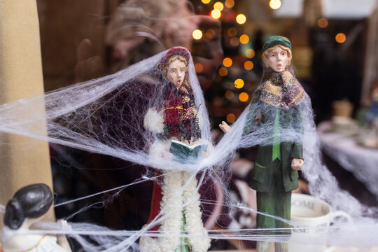 Two Victorian style caroler figurines covered in cobwebs stand near mugs and festive lights, evoking eerie nostalgia.